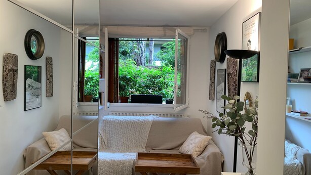 A cozy room with a sofa, a small table, and a window with a view of greenery. - Studette Meublée - Boulogne-billancourt Face au Stade de Roland-garros (Boulogne-Billancourt)