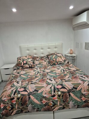 Ein Schlafzimmer mit einer gesteppten Kopfteile, einer zudeckenden Bettwäsche mit Blumenmuster, einem Nachttisch mit einer Dekoration und einer wandmontierten Klimaanlage.