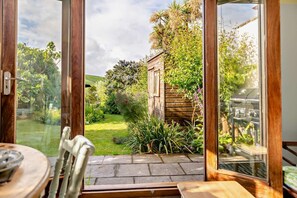 Une terrasse avec une table et des chaises, une plateforme en bois et une vue sur un jardin agrémenté d’un abri de jardin en bois et d’un barbecue.