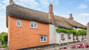 A traditional thatched-roof cottage with a stone facade, multiple windows, and a flower bed in the foreground.