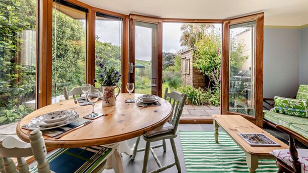 A dining area with a wooden table set for two, surrounded by chairs, with a view of a garden through large glass doors.