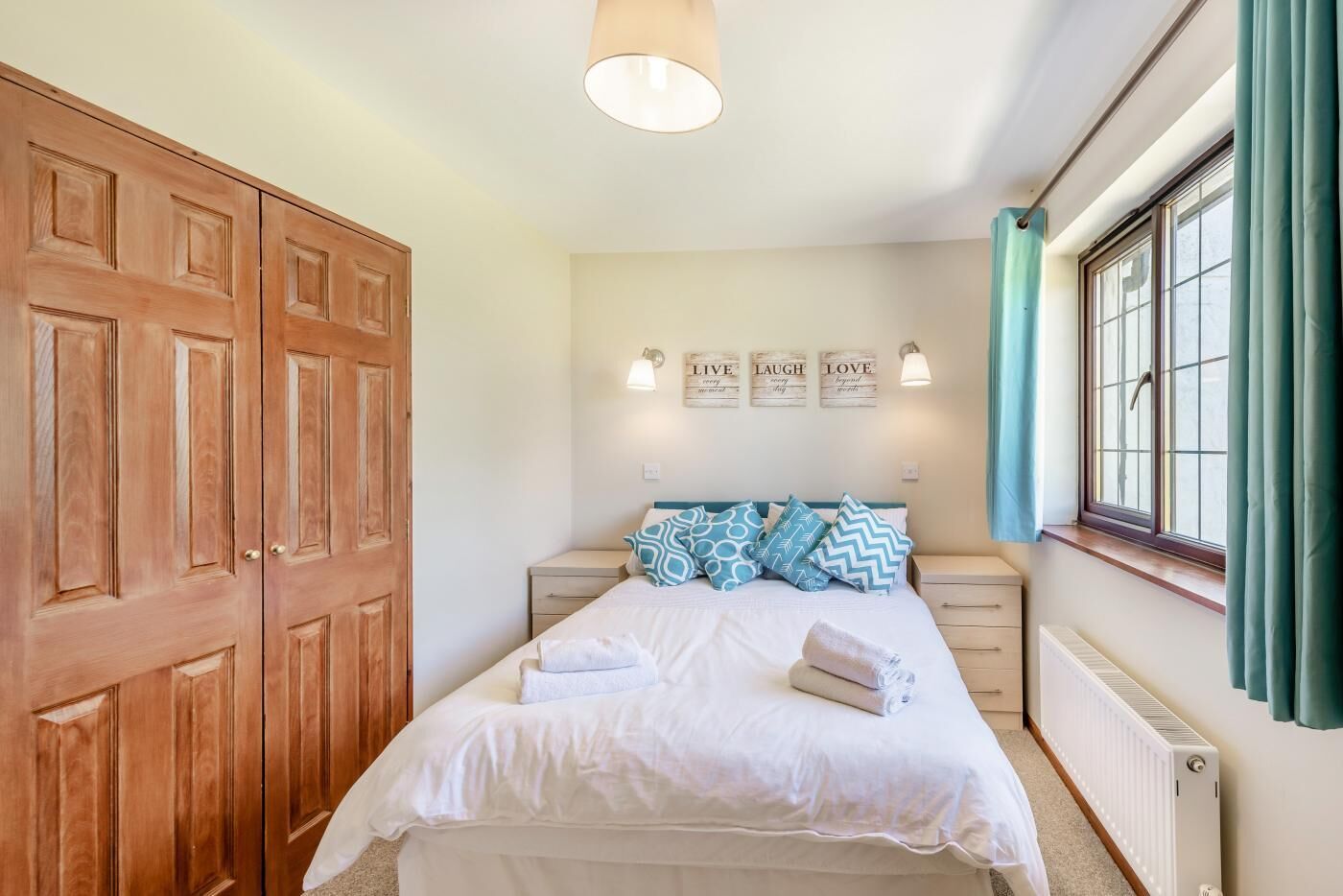 A bedroom with a bed, a wooden wardrobe, a nightstand, a wall-mounted light, a window with curtains, and a radiator.