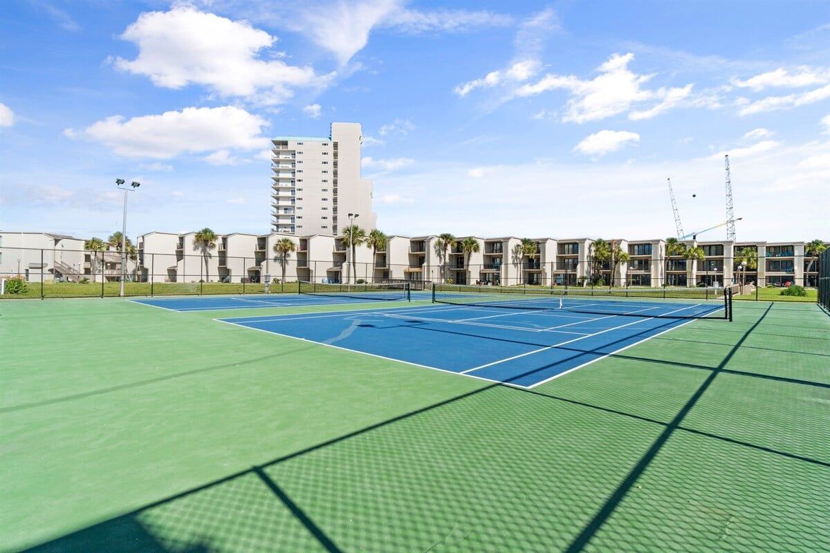 An outdoor tennis court with a net, surrounded by a fence, and adjacent to a building with multiple balconies.