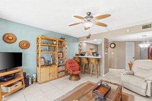 Un salon avec une télévision, des meubles en osier, un ventilateur de plafond et un coin cuisine à l’arrière-plan.