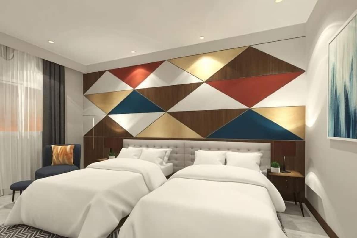A modern hotel room with a large bed, two bedside tables, a headboard with geometric patterns, and a large window with curtains.