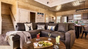 Un interior de cabaña acogedor con una escalera de madera, una sala de estar con sofá y mesa de centro, y una cocina con armarios y un refrigerador.