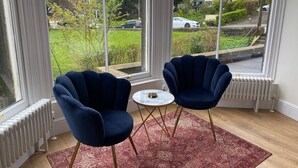 A room with two dark blue armchairs, a small round table, and a red patterned rug. Large windows offer a view of a green garden and a building with a dome. - The Matcham Studio in the heart of Buxton (Buxton)