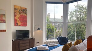 A hotel room with a large window, a bed with blue and white bedding, a television on a wooden cabinet, and a poster on the wall. - The Matcham Studio in the heart of Buxton (Buxton)