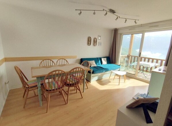A cozy living room with a wooden dining table and chairs, a sofa, and a balcony with a view.