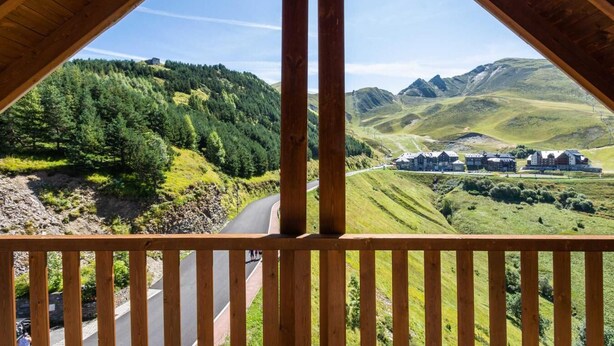 Een houten balkon met uitzicht op een kronkelende weg, weelderige groene heuvels en een groep gebouwen in de verte.