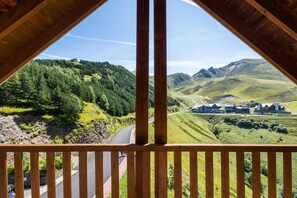 A wooden balcony with a view of a winding road, lush green hills, and a cluster of buildings in the distance.