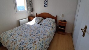Een slaapkamer met een bed, nachtkastje, lamp, raam met gordijnen en een kunstwerk aan de muur.