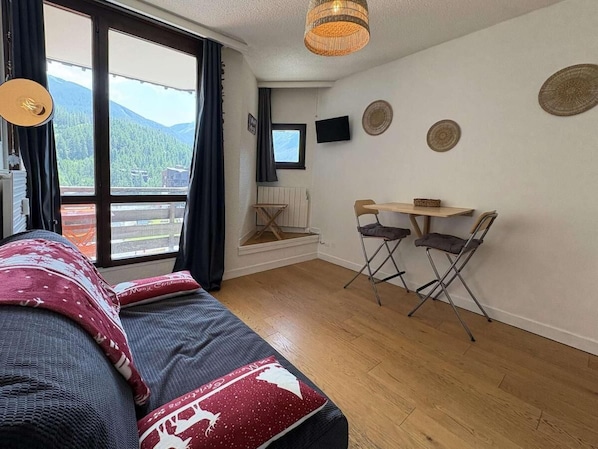 A bedroom with a bed, a desk, two chairs, a window with a view of mountains, and a television on the wall.