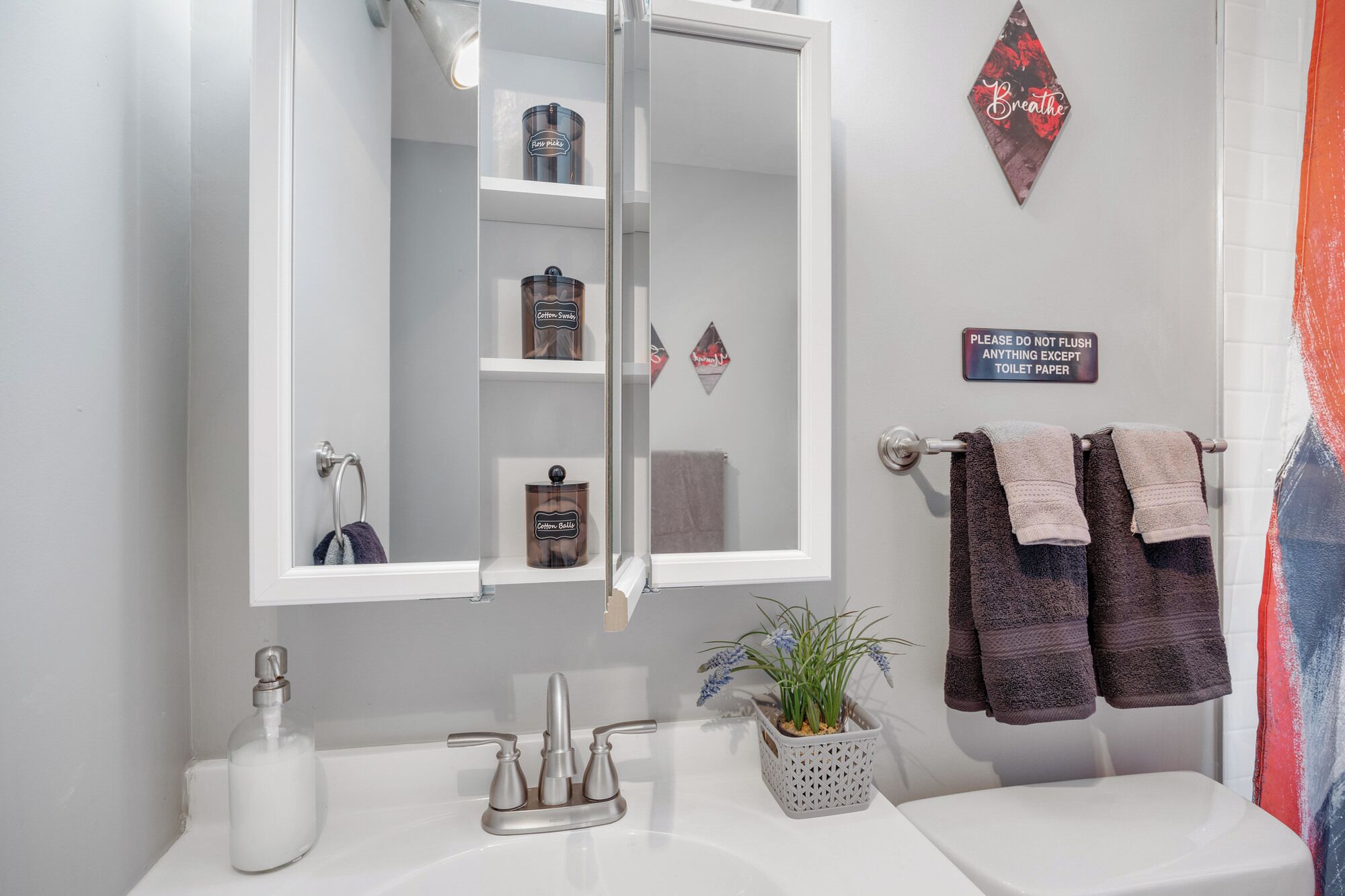 A bathroom with a white sink, a mirror, and a shelf with toiletry items. There is a sign on the wall and towels hanging on a rack.