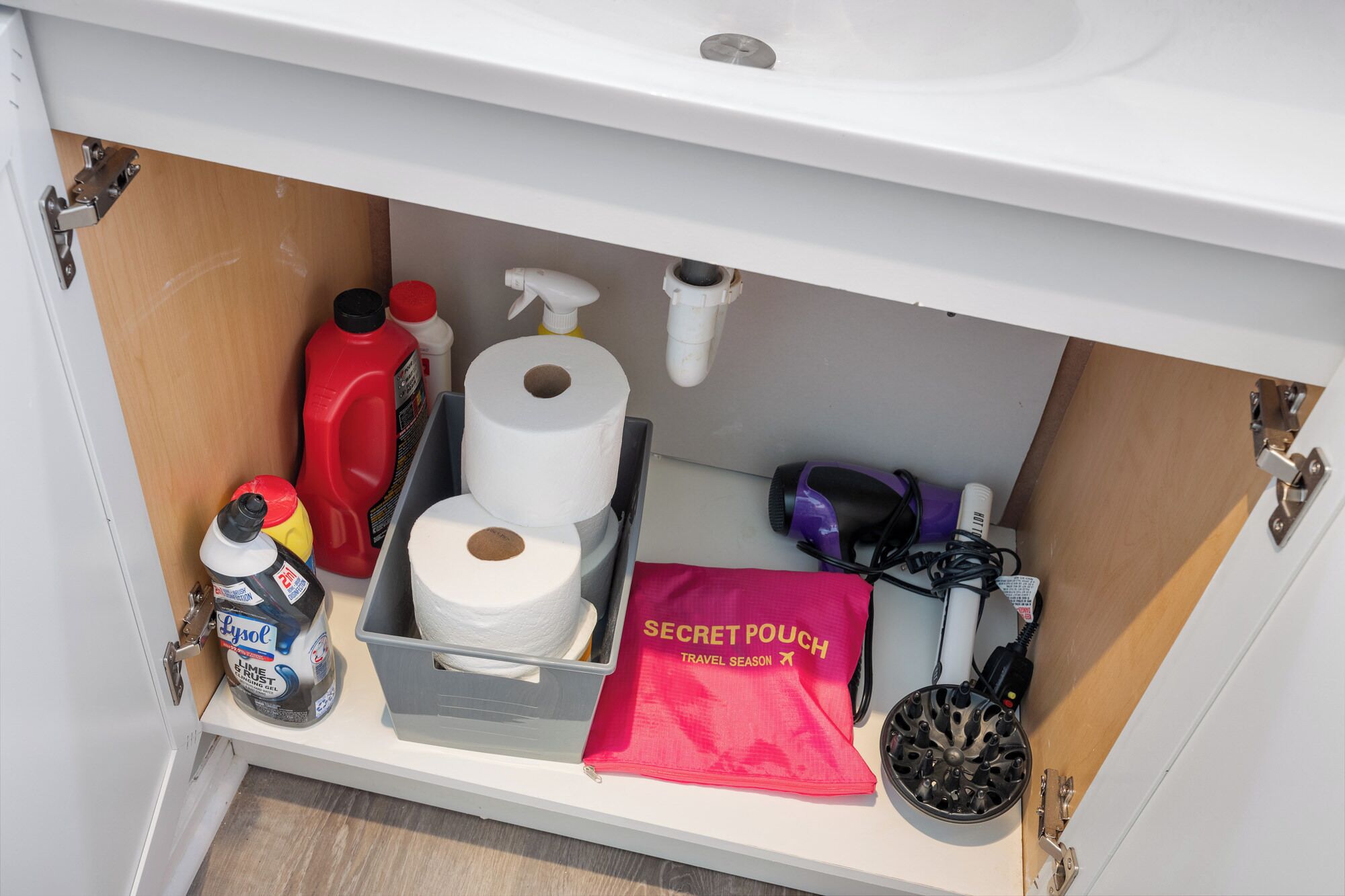 Un mueble de cocina con un estante que contiene una botella de Lysol, un rollo de papel higiénico, una bolsita roja etiquetada como “SECRET POUCH” y un secador de pelo.