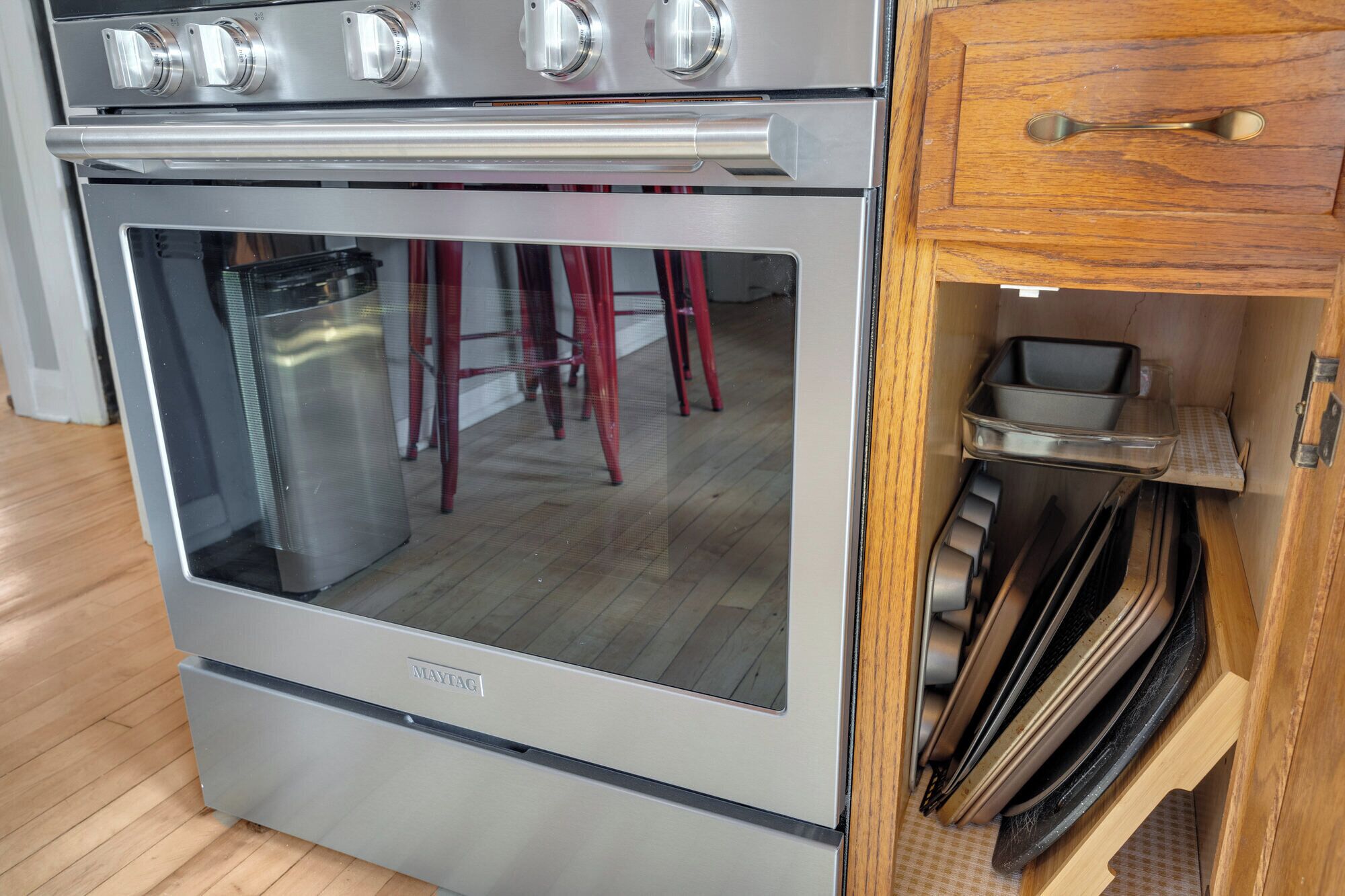 Un horno Maytag de acero inoxidable con puerta de vidrio, un gabinete de madera con asa metálica y una bandeja de metal en su interior.
