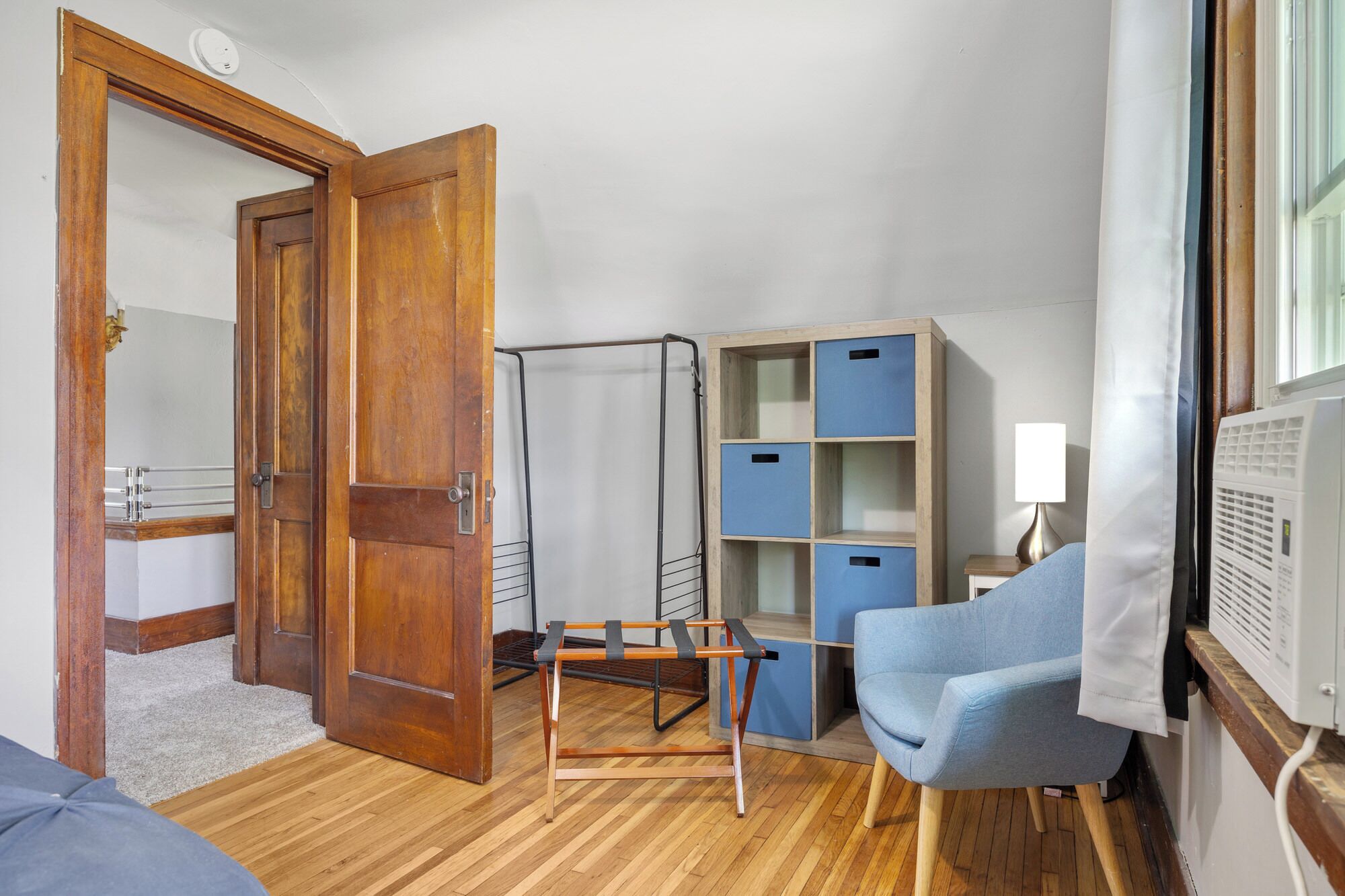 Una habitación con puerta de madera, un perchero metálico, una silla azul, una lámpara blanca y un aire acondicionado blanco.
