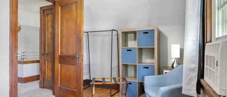 Una habitación con puerta de madera, un perchero metálico, una silla azul, una lámpara blanca y un aire acondicionado blanco.