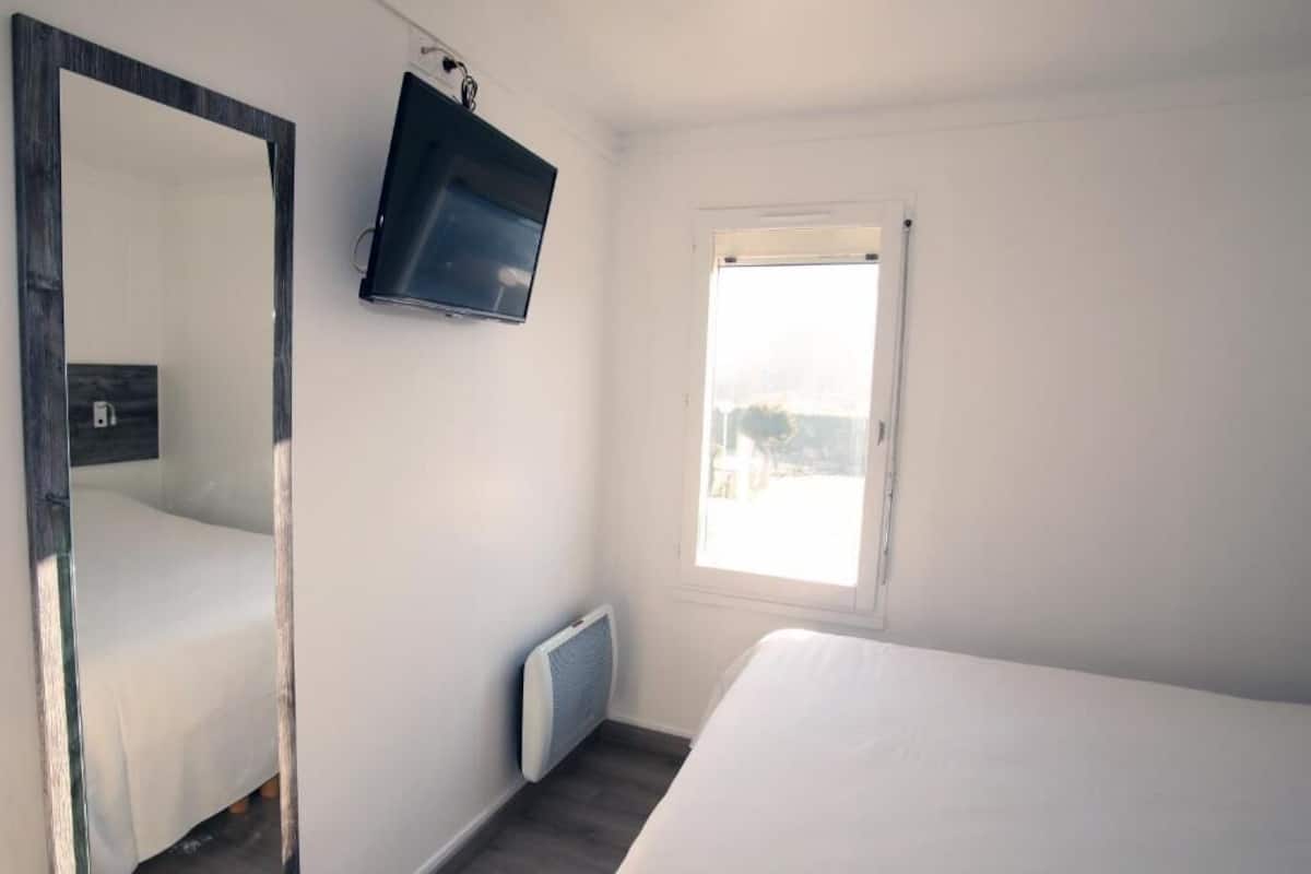 A bedroom with a bed, a large mirror, a TV mounted on the wall, and a window with a view of the outdoors.