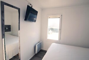 Une chambre à coucher avec un lit, un grand miroir, un téléviseur fixé au mur et une fenêtre donnant sur l’extérieur.