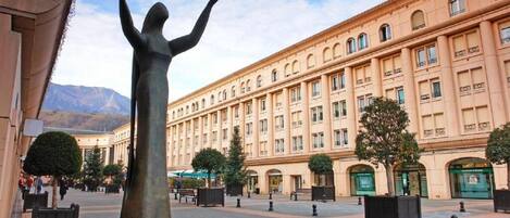 Una plaza pública con una estatua, un edificio grande con muchas ventanas y un cielo despejado.
