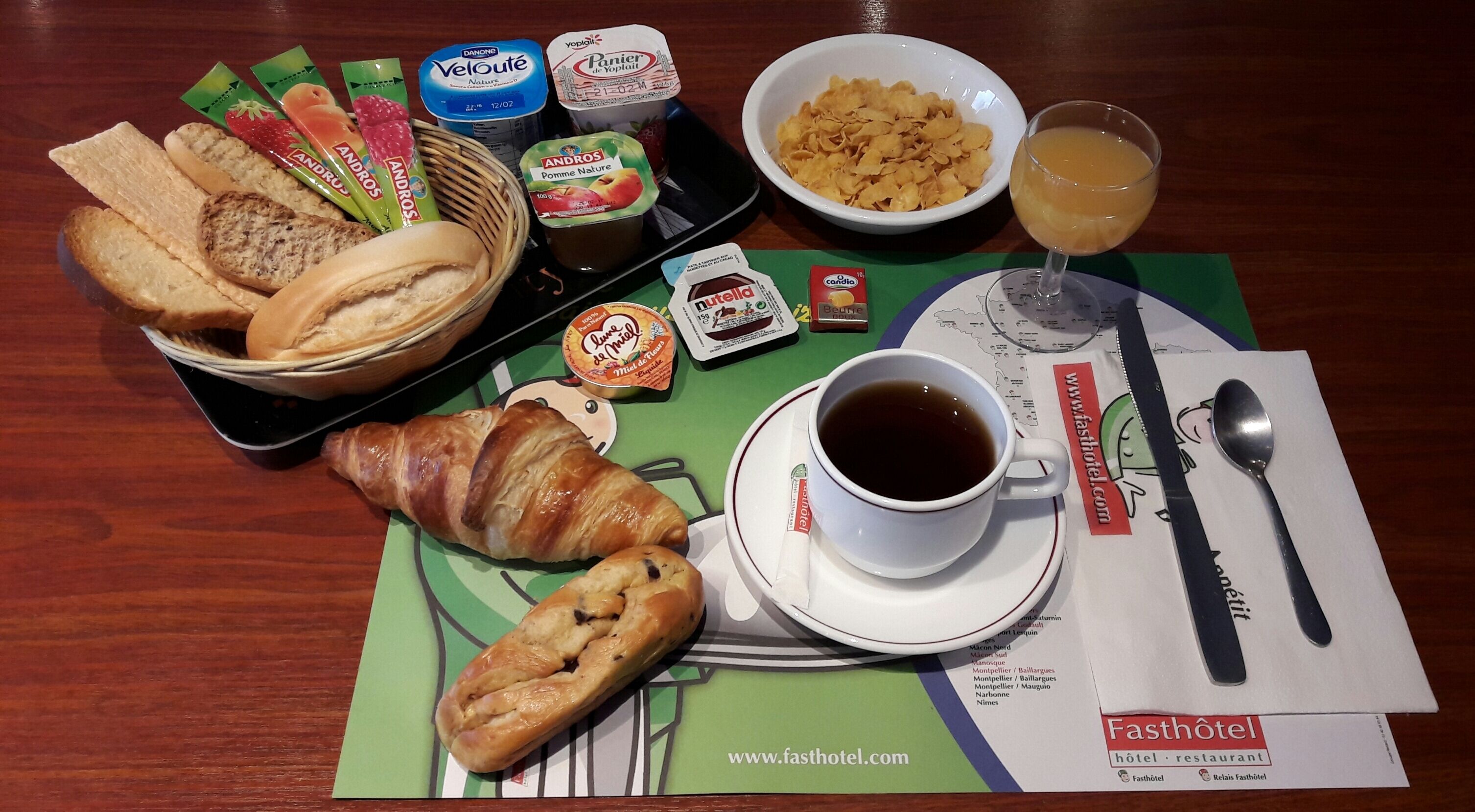 A breakfast setting with bread, pastries, yogurt, cereal, and a cup of coffee on a table with a placemat.