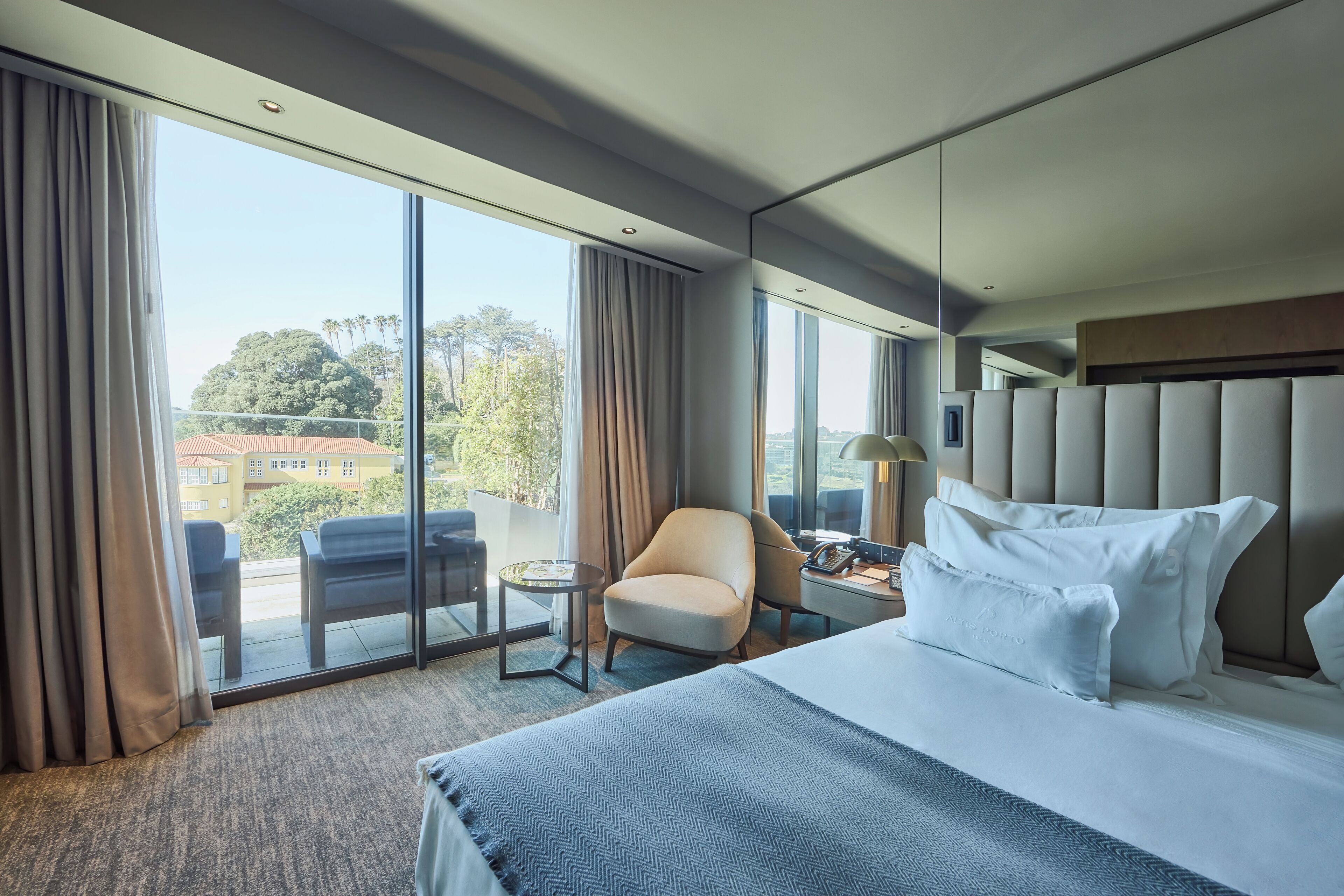 A modern hotel room with a large bed, a chair, a small table, and a view of the city through large windows.
