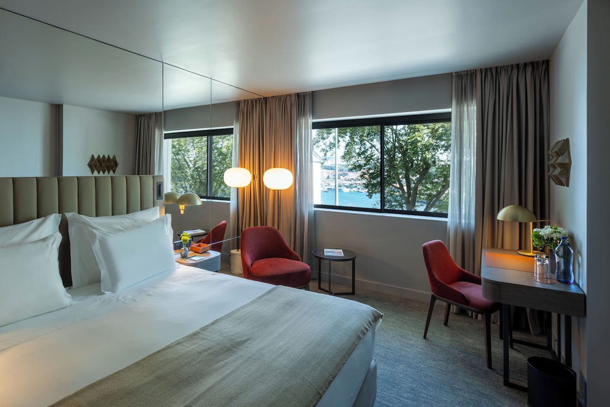 A hotel room with a large bed, two red chairs, a small table, and a desk with a lamp.