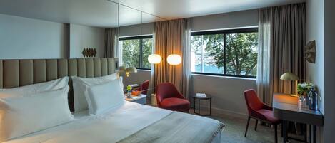 A hotel room with a large bed, two red chairs, a small table, and a desk with a lamp.