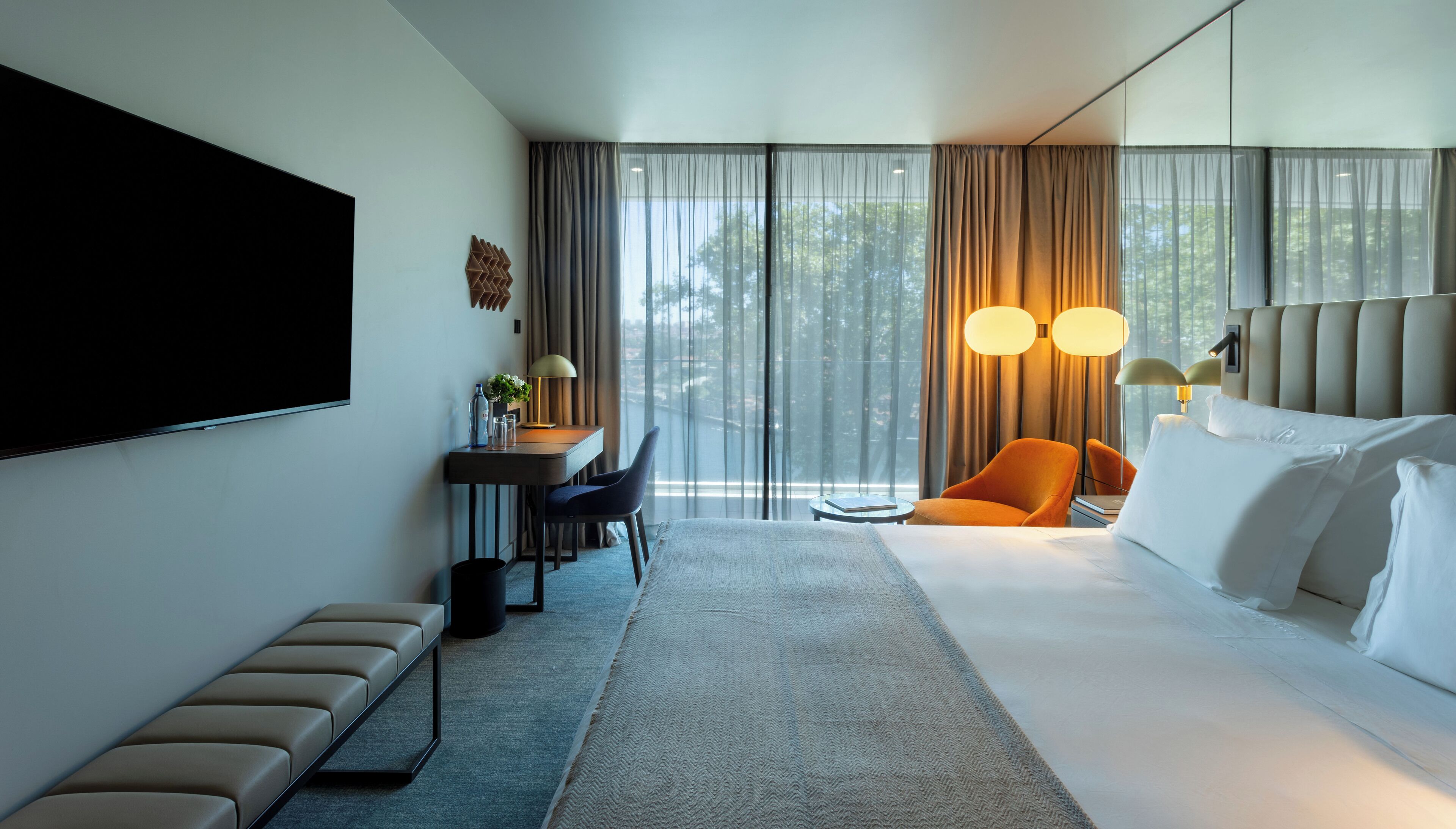 Une chambre d’hôtel moderne avec un grand lit, un bureau avec une chaise, une télévision et une vue sur les arbres par la fenêtre.