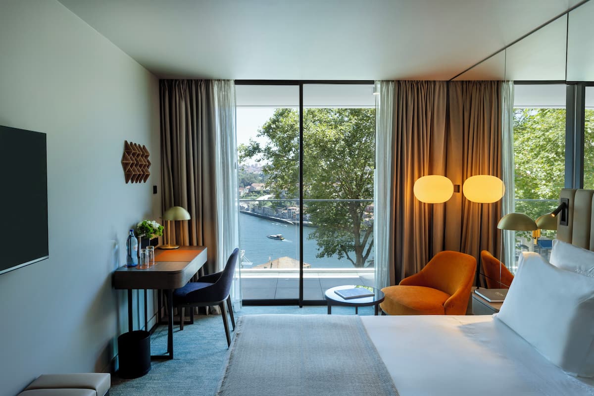 A modern hotel room with a large bed, a desk with a chair, a small table, and a view of a river and cityscape through large windows.
