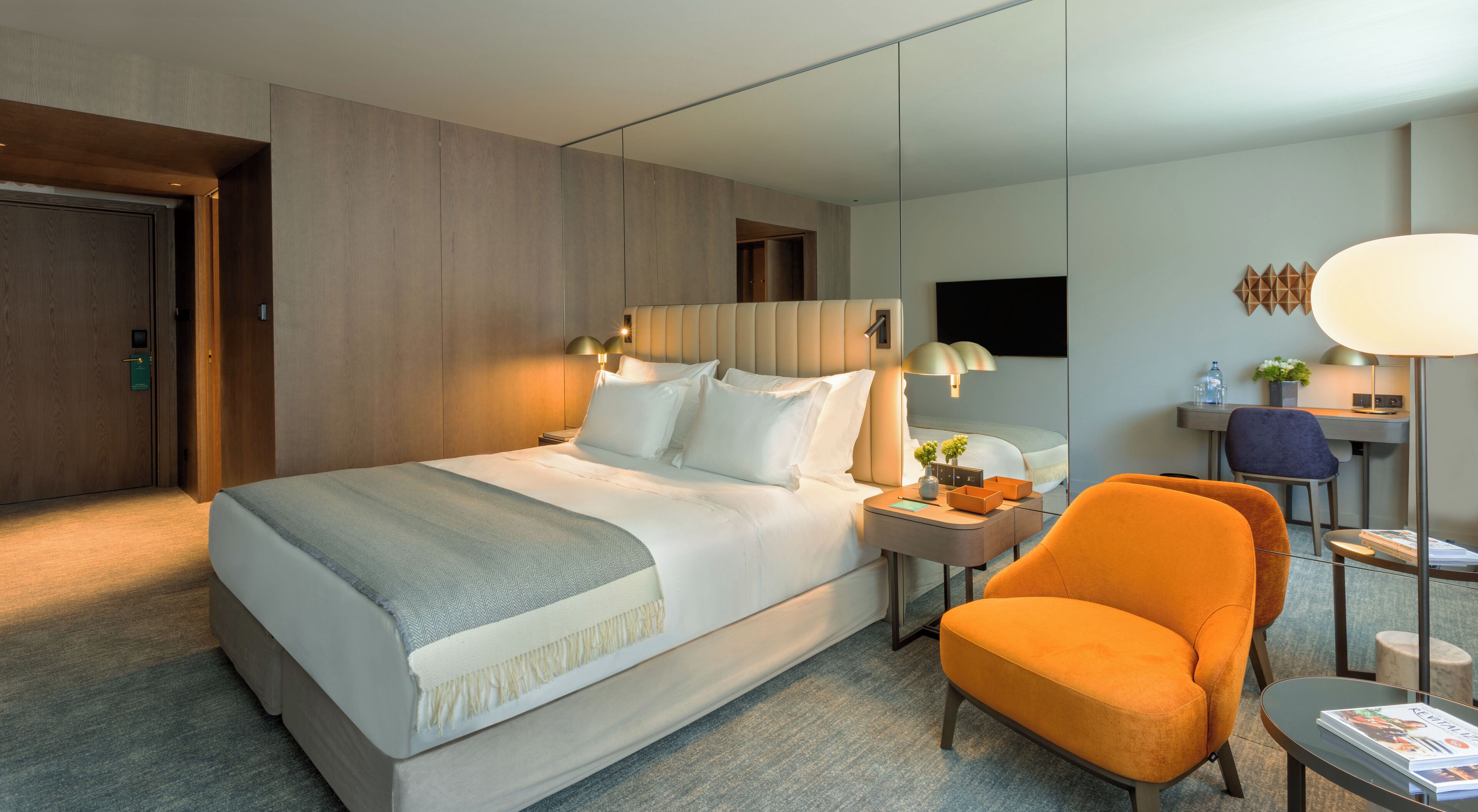 Une chambre d’hôtel moderne avec un grand lit, un bureau avec une chaise et un coin salon avec un fauteuil orange.