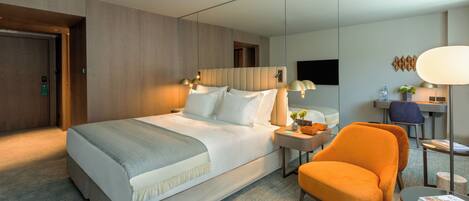 Une chambre d’hôtel moderne avec un grand lit, un bureau avec une chaise et un coin salon avec un fauteuil orange.