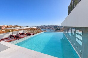 Una piscina infinita en la azotea con sillas reclinables y sombrillas rojas, con vistas a un paisaje urbano y un puerto.