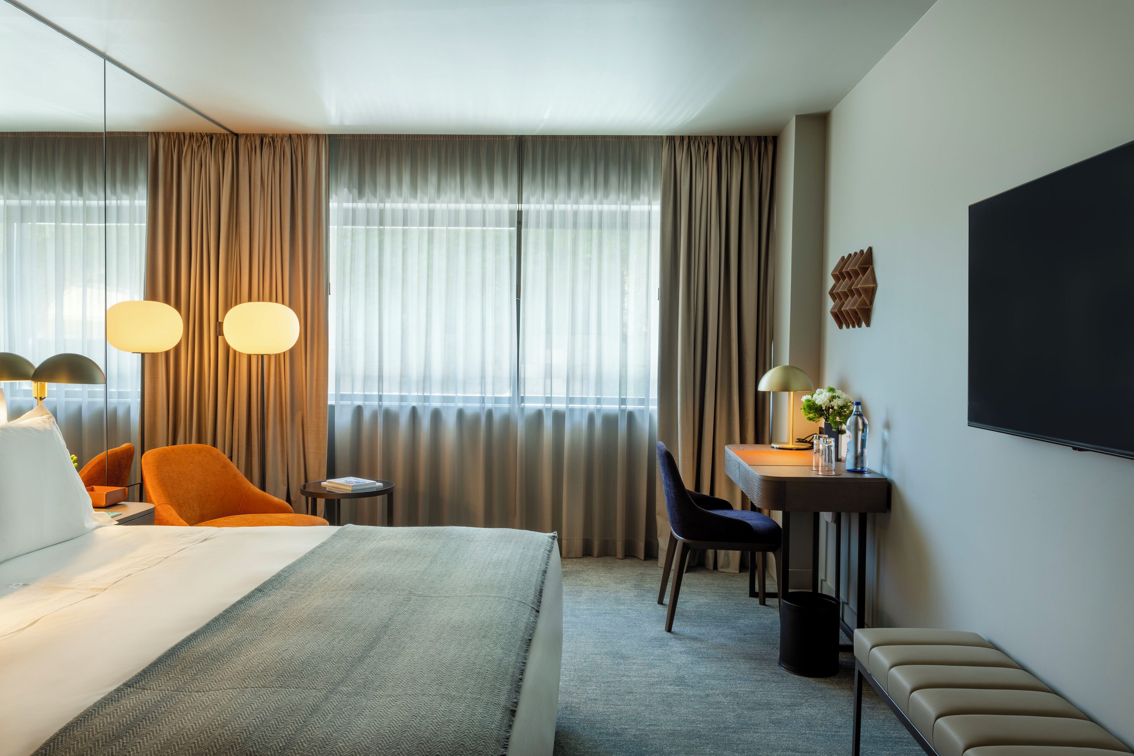 A hotel room with a large bed, two chairs, a desk with a lamp, and a TV on the wall.