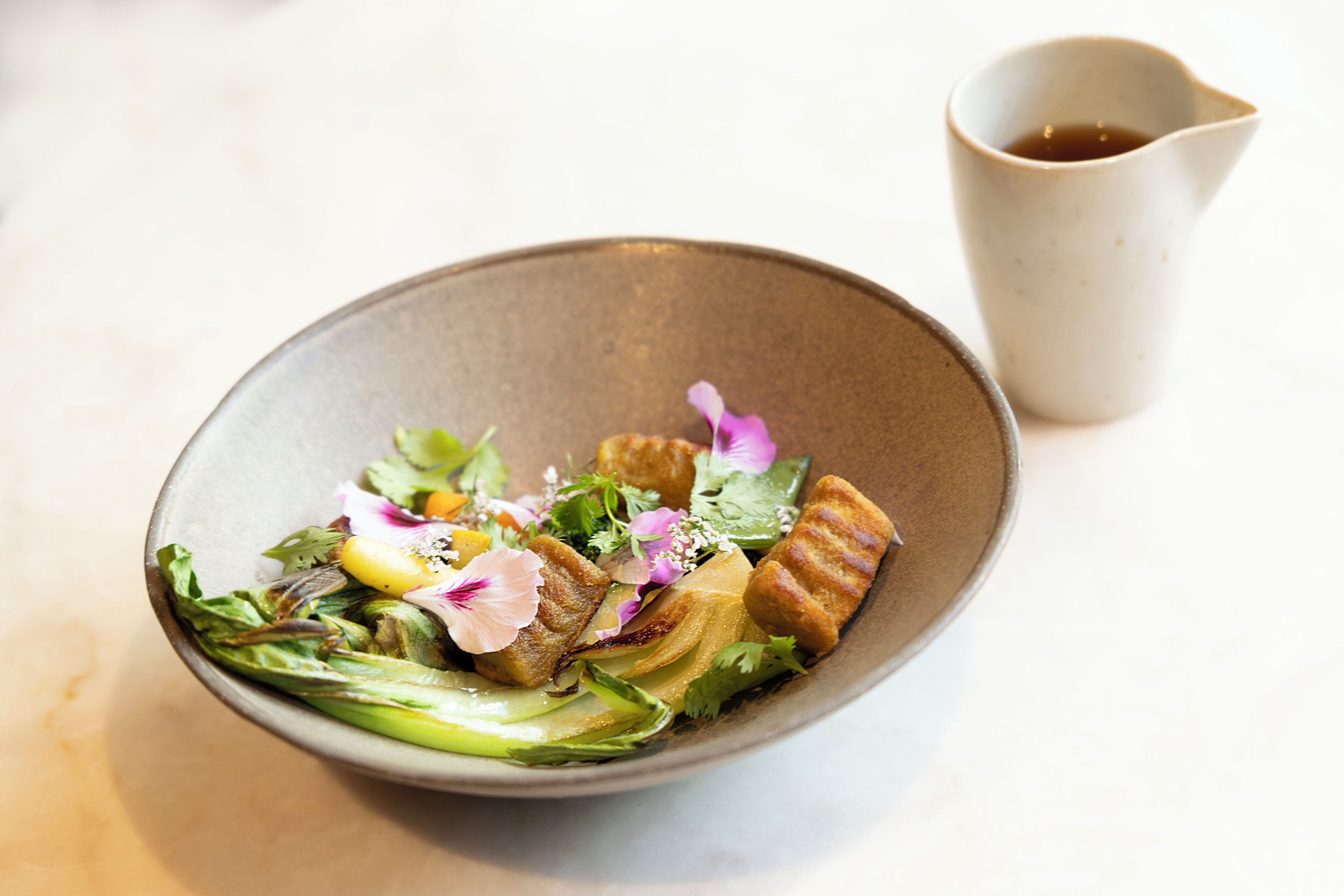 A bowl of salad with various greens and vegetables, accompanied by a small pitcher of sauce.