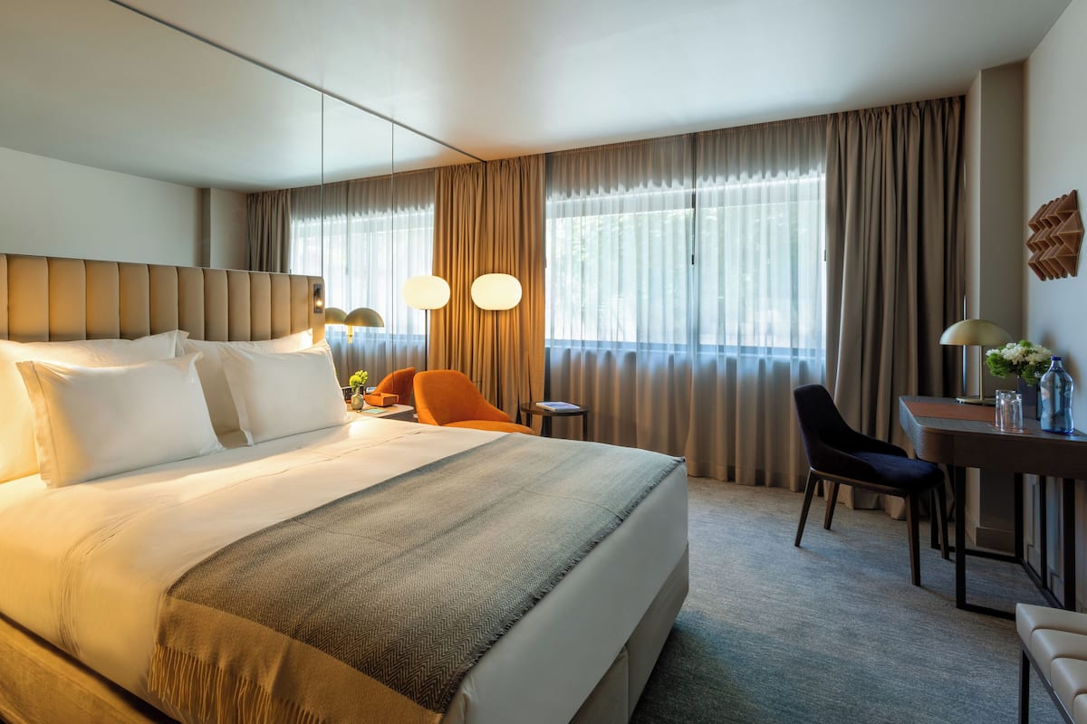 A hotel room with a large bed, a desk, a chair, and a nightstand with a lamp.
