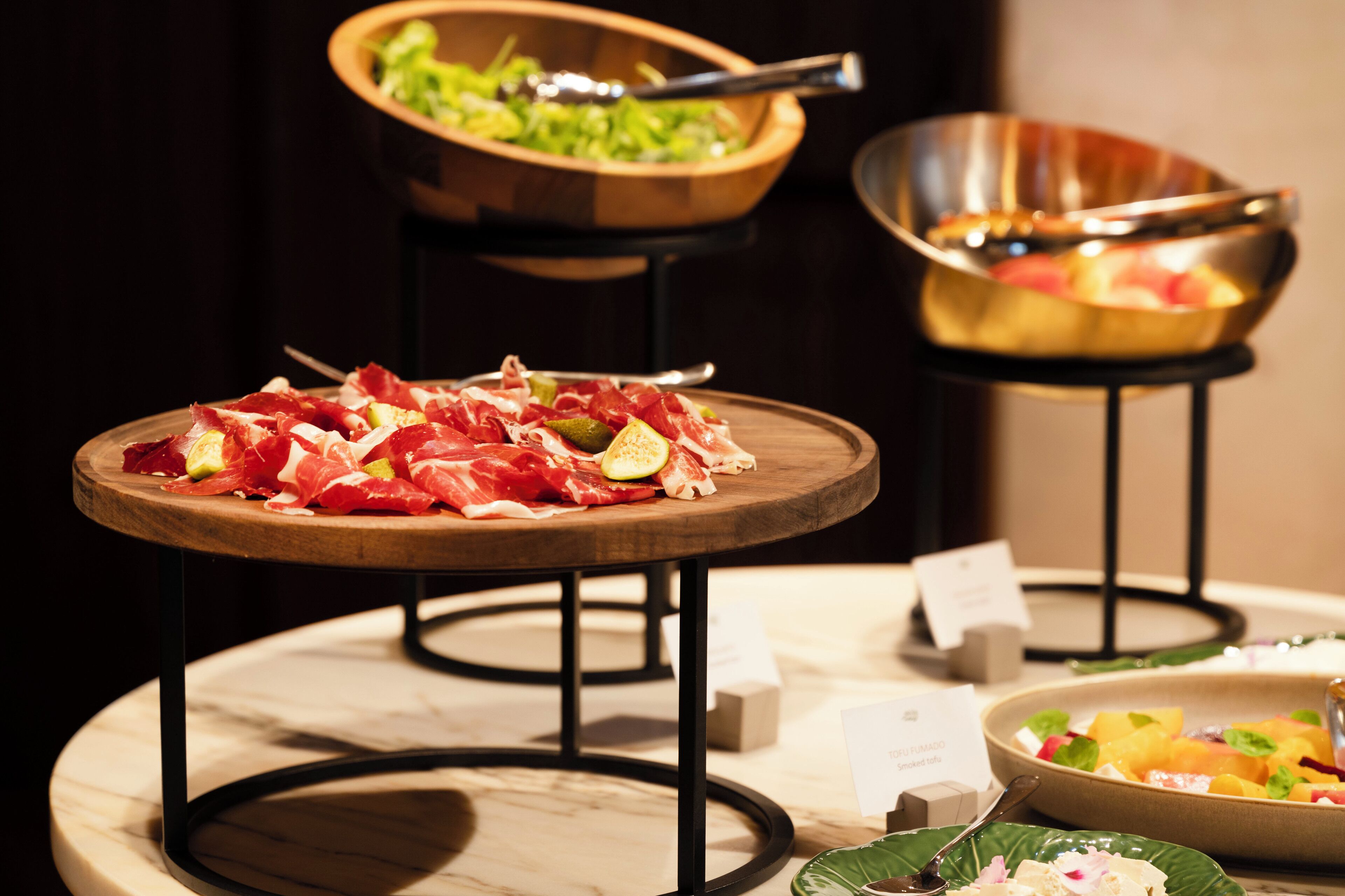 A dining area with a wooden table displaying various meats and vegetables, a bowl of salad, and a bowl of fruit on a marble table.