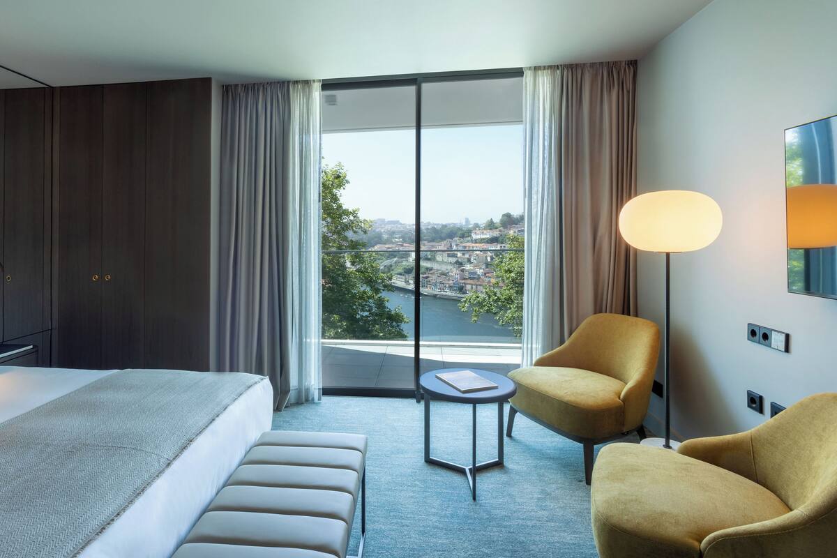 A modern hotel room with a large bed, a small round table, a sofa, and a view of a cityscape through large windows.