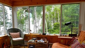 Ein gemütliches Blockhaus-Interieur mit Holzboden, einem gepolsterten Sessel, einer Couch und einem Tisch. Große Fenster bieten Blick auf einen bewaldeten Bereich.