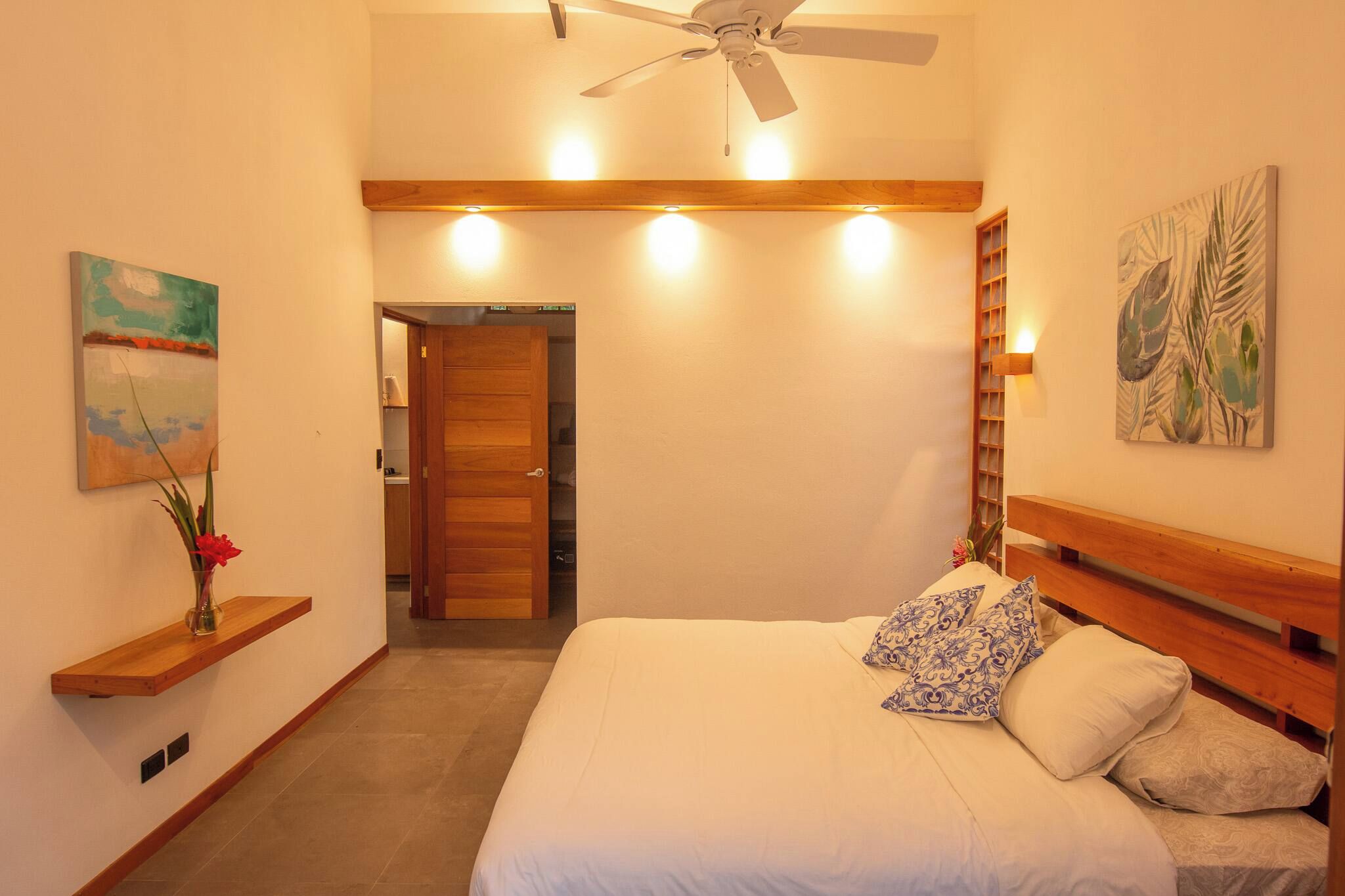 A bedroom with a bed, a ceiling fan, a wooden headboard, a nightstand, a wall-mounted light, and a door leading to a bathroom.