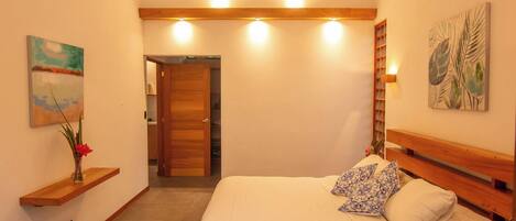 A bedroom with a bed, a ceiling fan, a wooden headboard, a nightstand, a wall-mounted light, and a door leading to a bathroom.