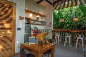 A modern kitchen with wooden cabinets, a refrigerator, and a coffee machine. There is a dining area with a wooden table, chairs, and a fruit bowl. The room has a large window with a view of tropical plants.