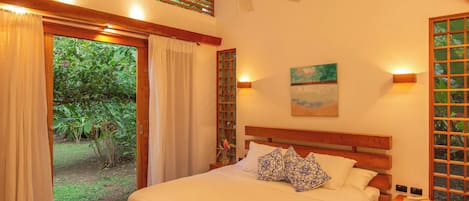 A bedroom with a bed, a ceiling fan, a painting, and a view of greenery outside.