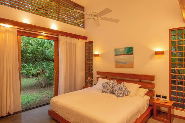 A bedroom with a bed, a ceiling fan, a painting, and a view of greenery outside.