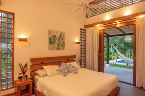 Ein Schlafzimmer mit einem Bett, einem Nachttisch, einer Wandlampe, einem Deckenventilator und Blick auf einen Garten mit einer Hängematte.