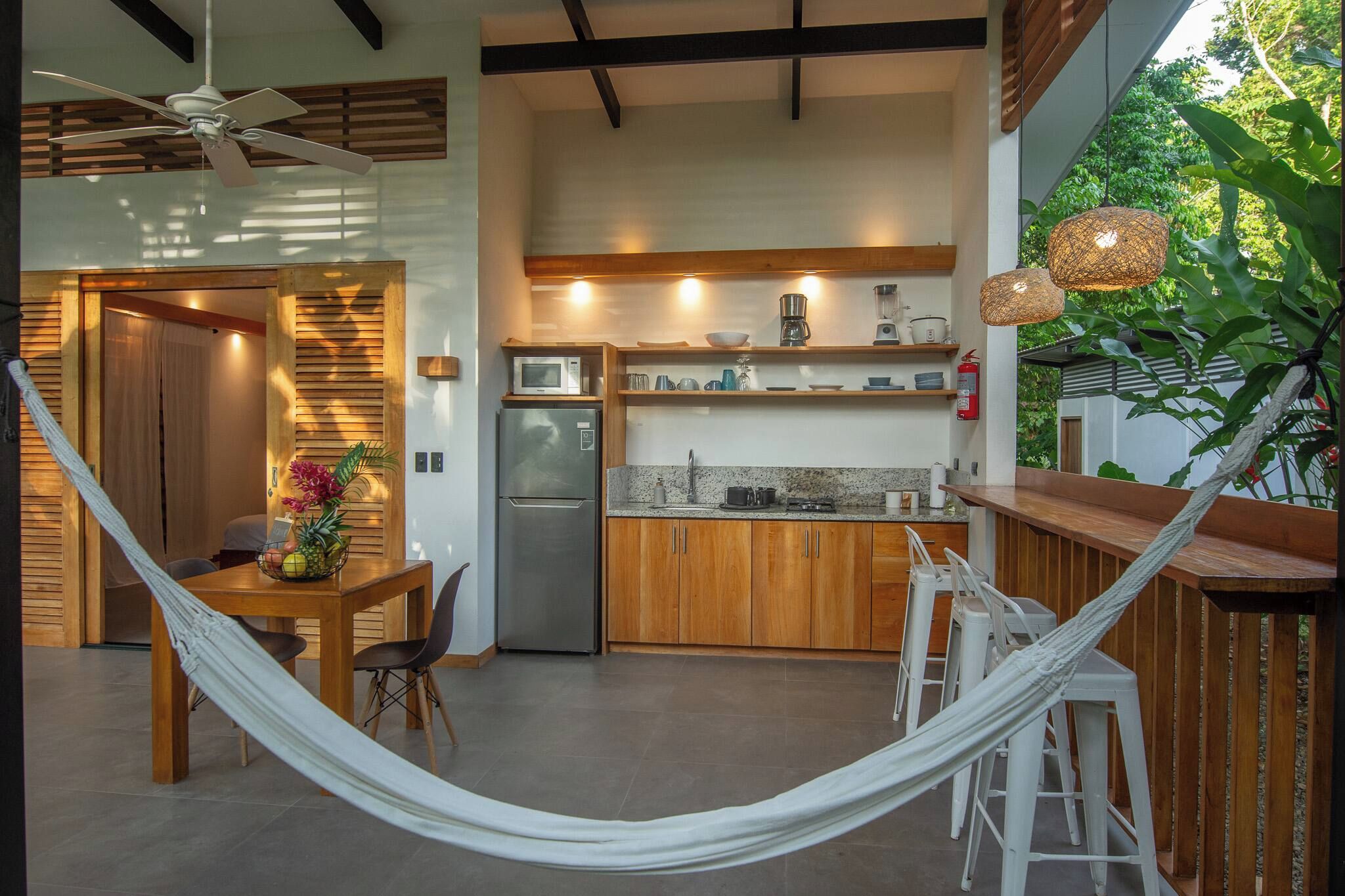 A modern kitchen with a freestanding refrigerator, a microwave, and a coffee maker on the countertop. There is a dining area with a table and chairs, and a hammock in the foreground.