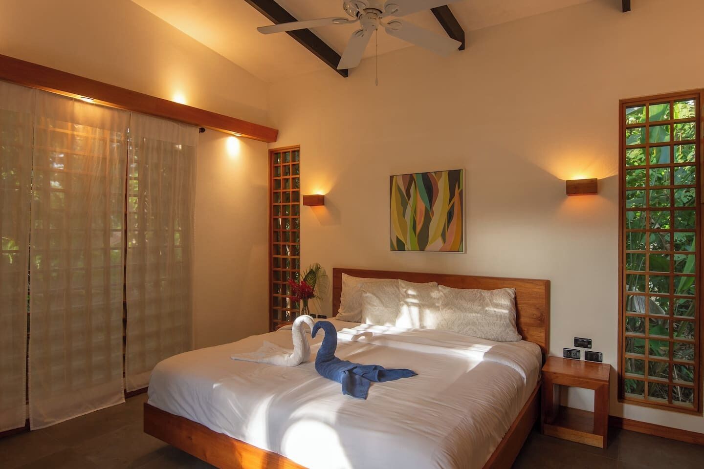 A hotel room with a bed, a ceiling fan, a window with a view of greenery, and a wall-mounted artwork.