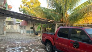 Een rode pick-up truck staat geparkeerd op een betegeld terras, met een palmboom en een gebouw op de achtergrond.