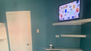 Een kamer met turquoise wanden, een witte deur en een betegelde vloer. Een televisie op een plank toont kleurrijke objecten. Twee zwarte barkrukken staan tegen de muur.